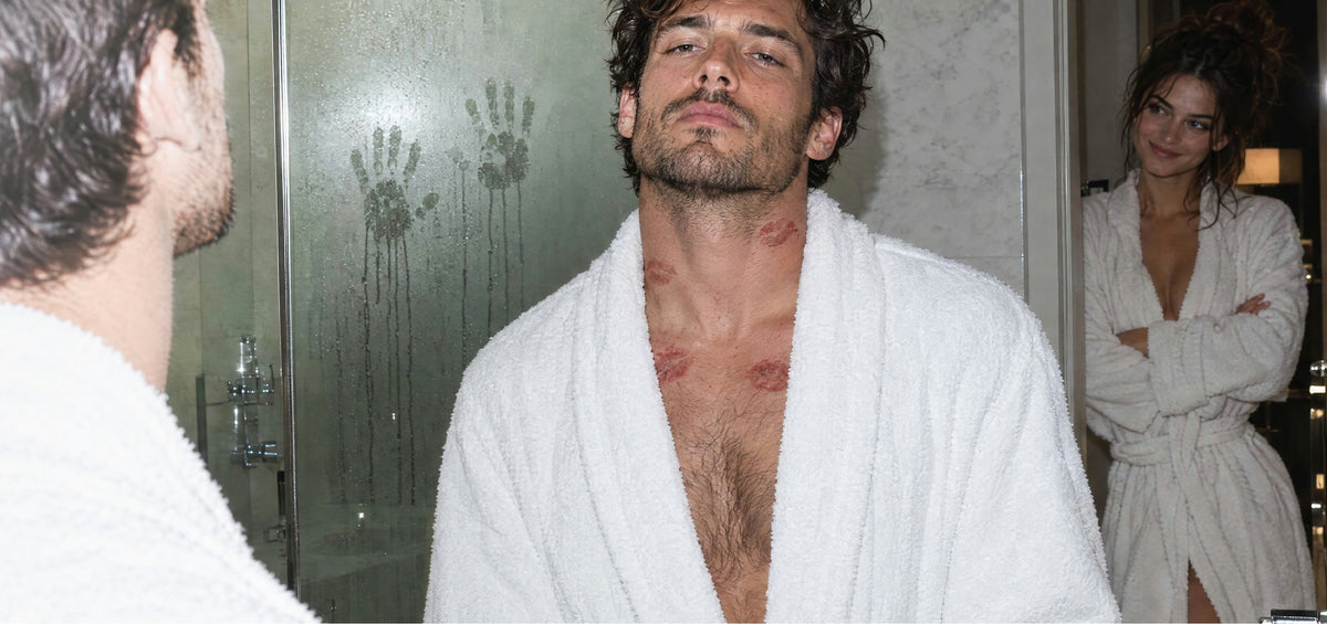 Man in a white bathrobe with lipstick kisses on his neck, a woman smiling in the doorway behind him